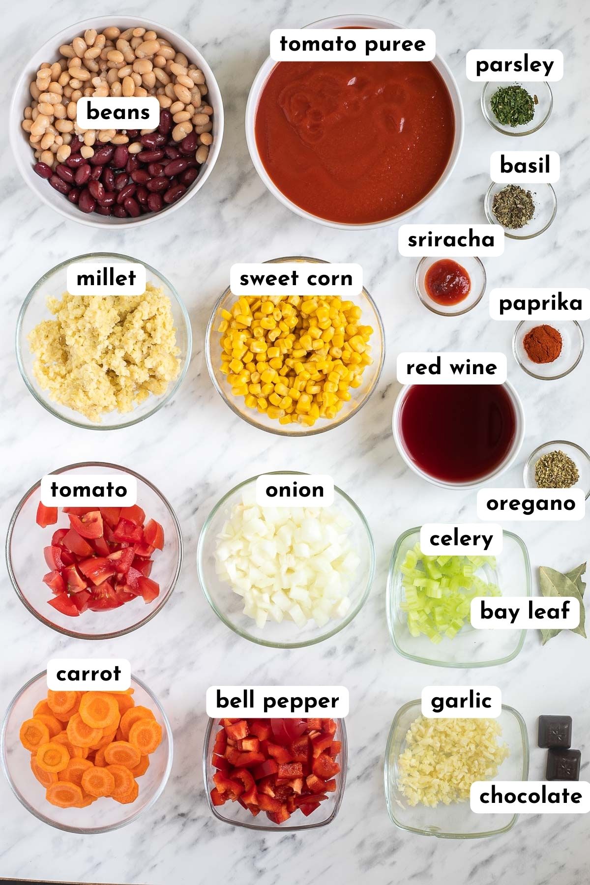 Ingredients of vegan chili in small bowls like beans, corn, millet, tomato puree, chopped tomato, onion, garlic, carrot, red pepper, red wine and different spices.