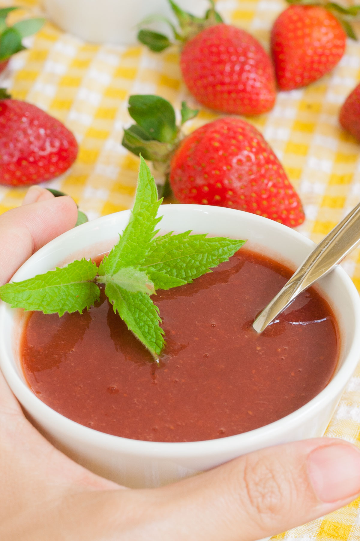A hand is holding a small white bowl with strawberry puree and some mint leaves. More fresh strawberries are scattered around it on a yellow tablecloth.