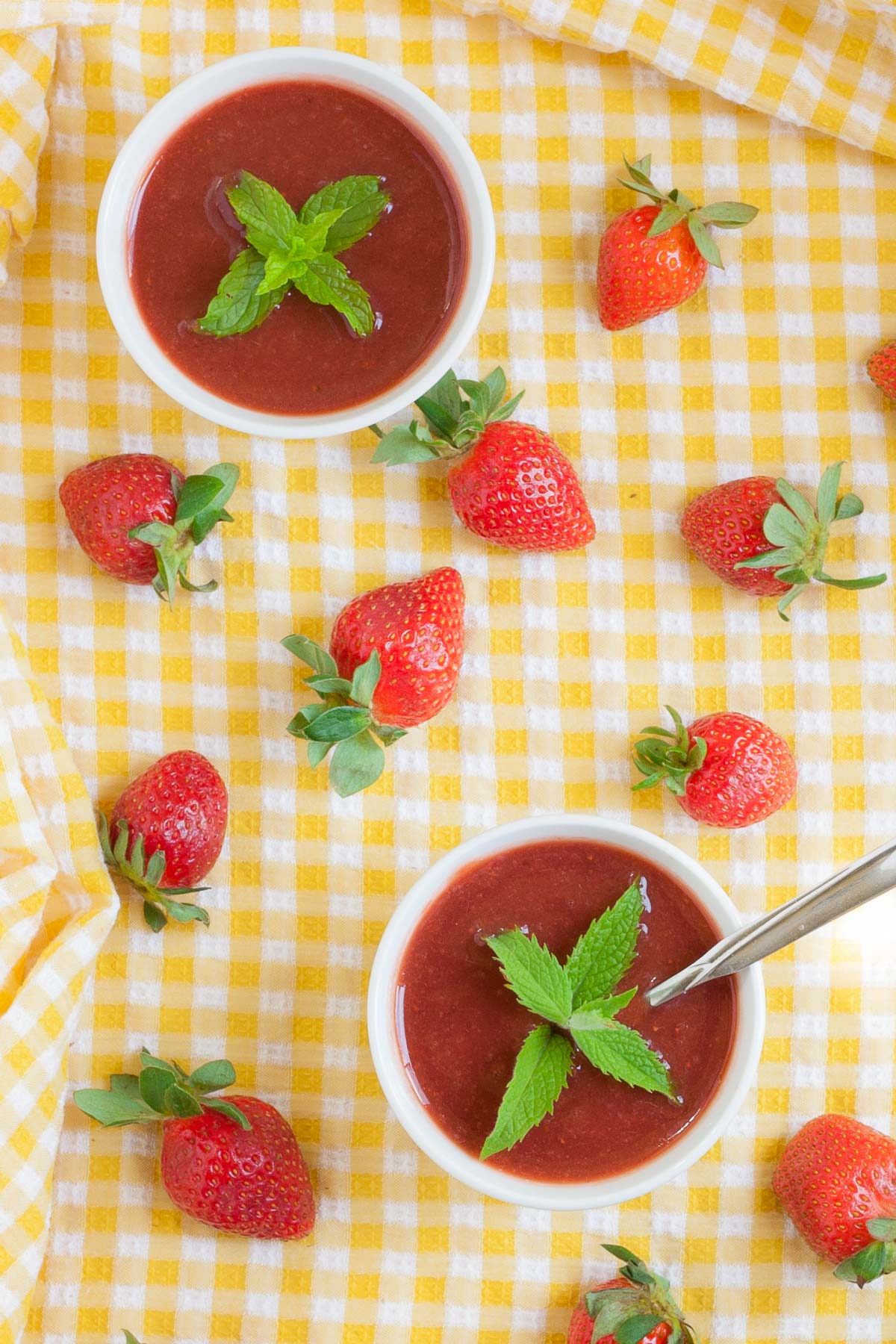 Two small white bowls with strawberry puree and some mint leaves. More fresh strawberries are scattered around it on a yellow tablecloth.