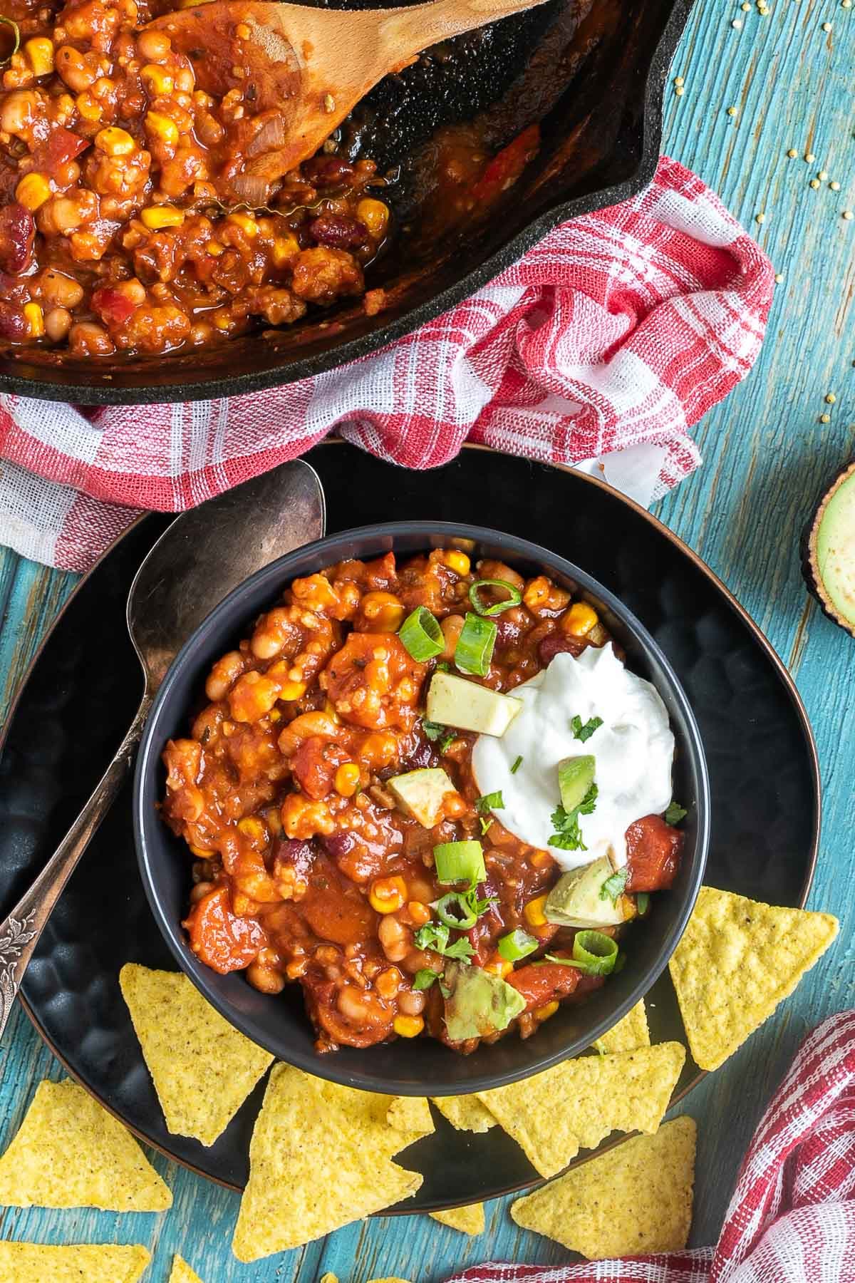 A black bowl with a chunky chili loaded with chopped vegetables and beans, topped with white cream, chopped avocado, and scallion. Leftovers are in a cast iron skillet next to it.