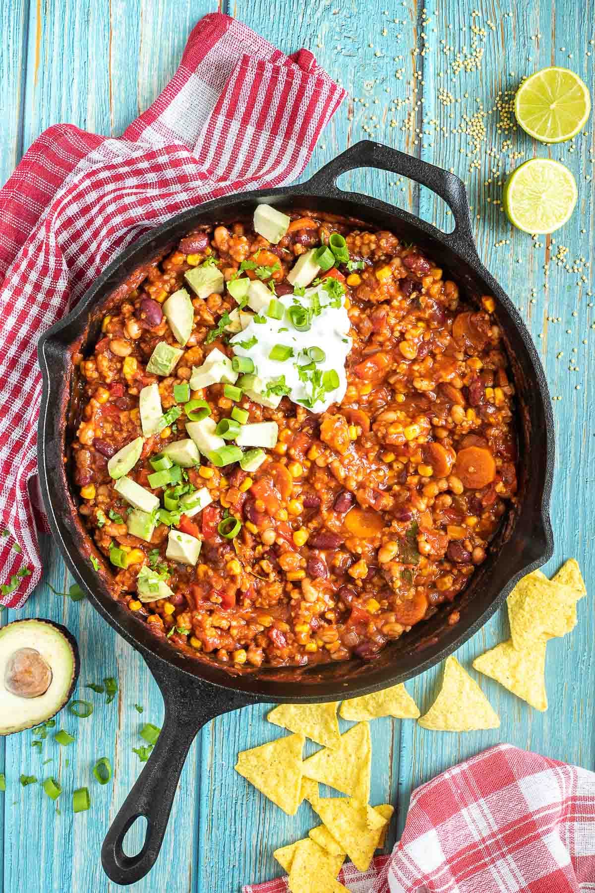 Cast iron skillet with a chunky chili loaded with chopped vegetables and beans, topped with white cream, chopped avocado, and scallion. Half avocados and tortilla chips are around it.