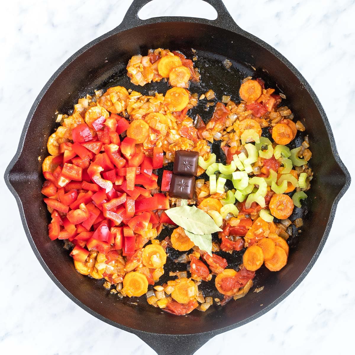 Cast iron skillet with chopped onion, sliced carrots, chopped tomatoes, celery, chocolate cubes, bay leaves, and chopped red peppers.