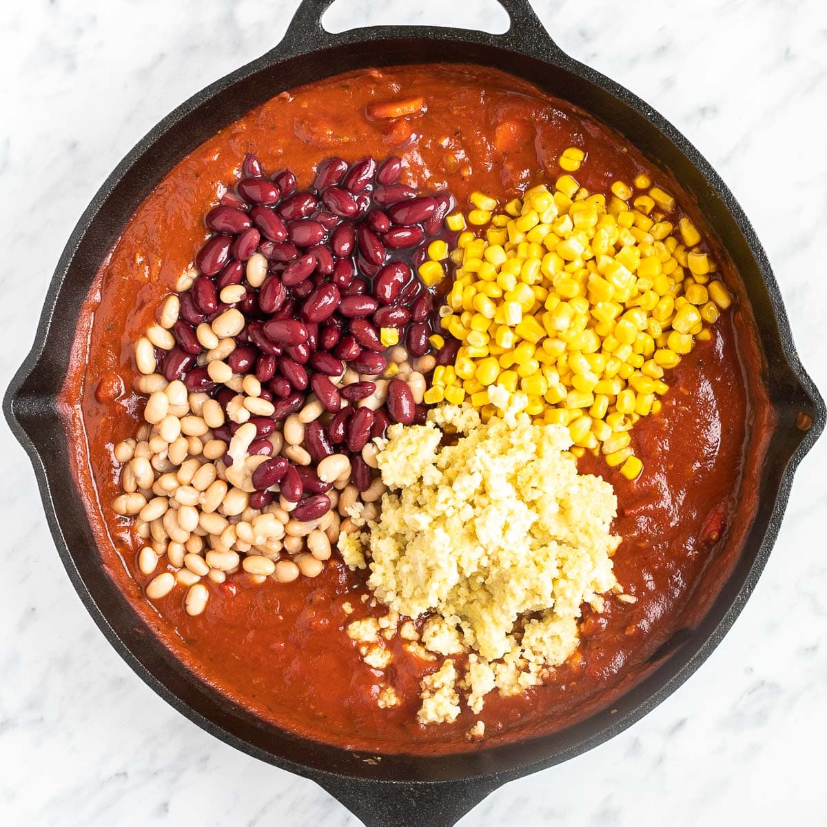 Cast iron skillet with chunky tomato sauce topped with red kidney beans, white beans, sweet corn, and cooked millet.