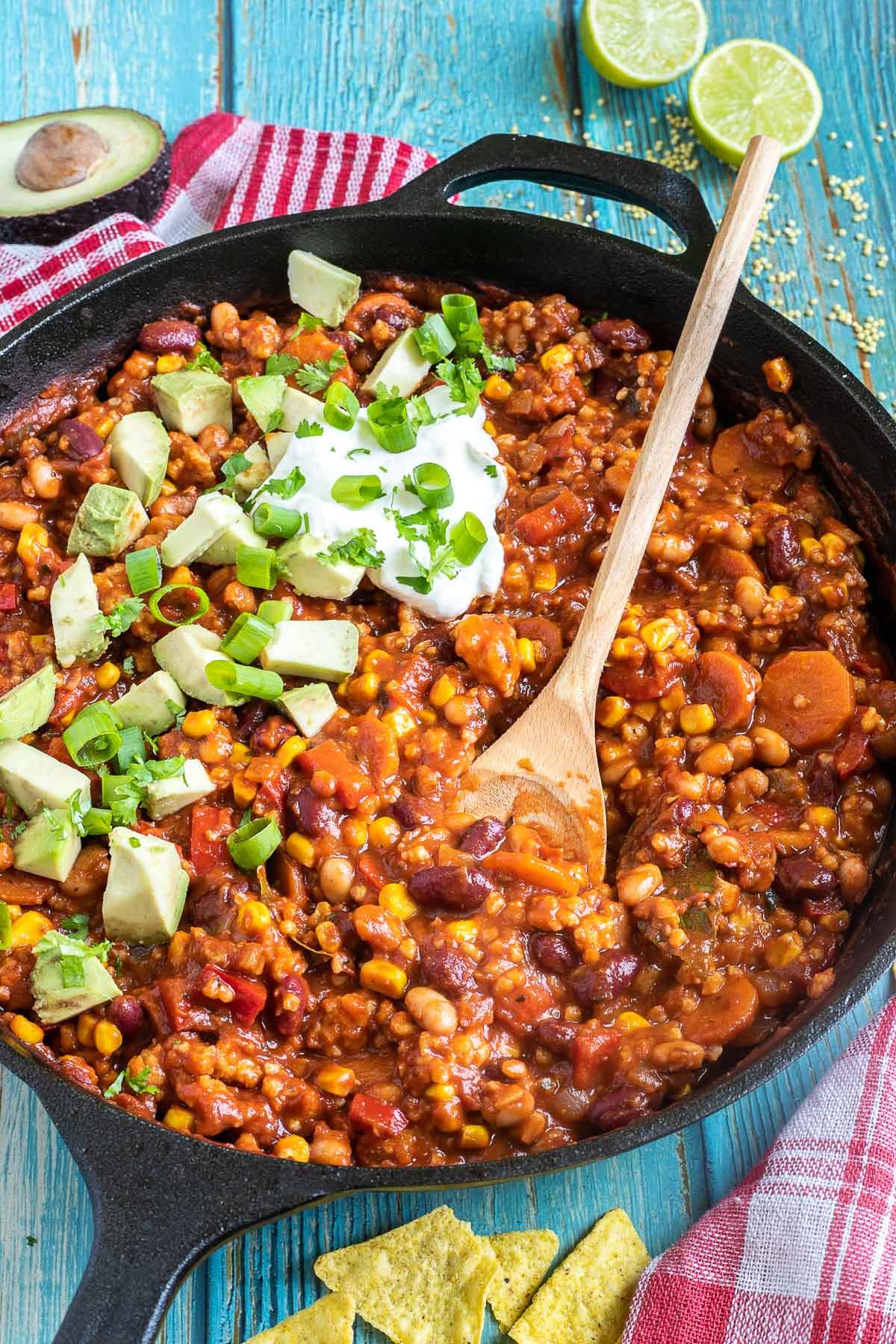 Cast iron skillet with a chunky chili loaded with chopped vegetables and beans, topped with white cream, chopped avocado, and scallion. A wooden spatula is placed in the middle.