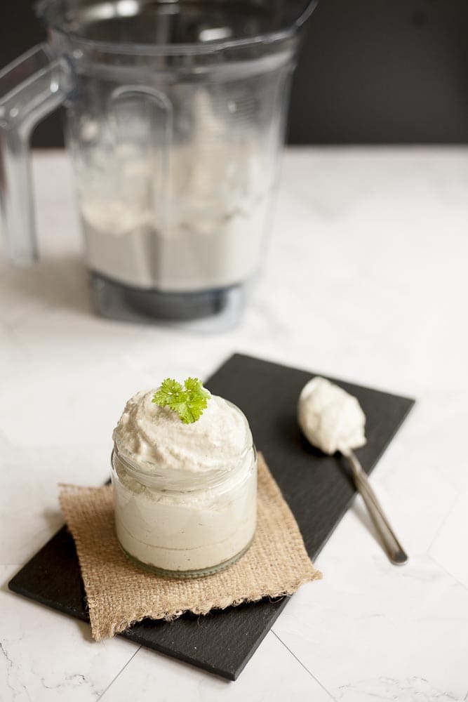 Small white glass packed with dairy-free sour cream and topped with fresh parsley. At the background the blender with the remaining sour cream. 