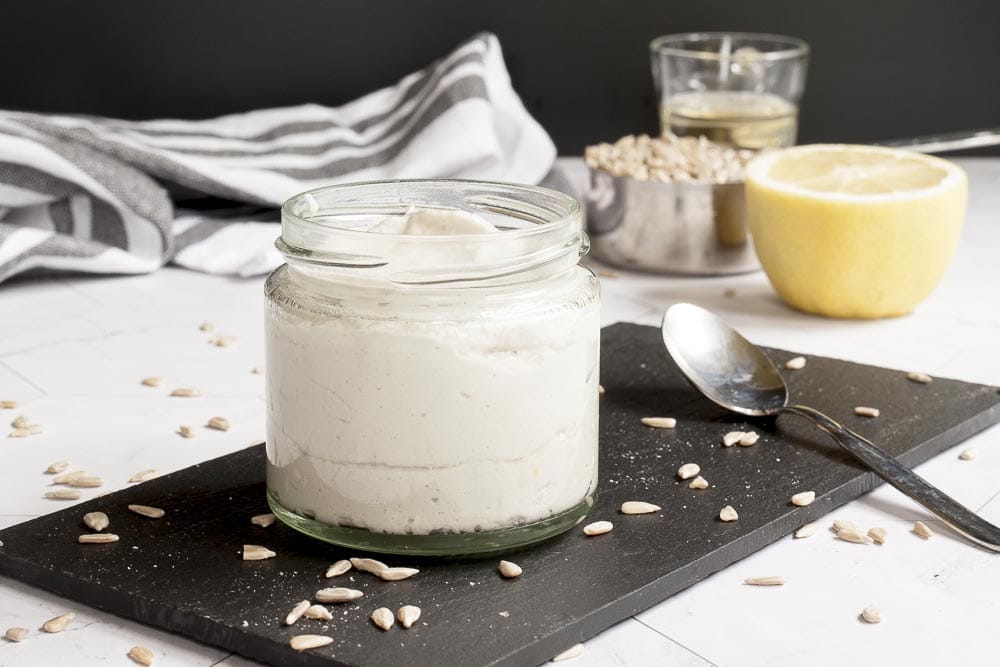 Small white glass packed with dairy-free sour cream. At the background the ingredients: a cup of sunflower seed, half of lemon and a small glass of apple cider vinegar 