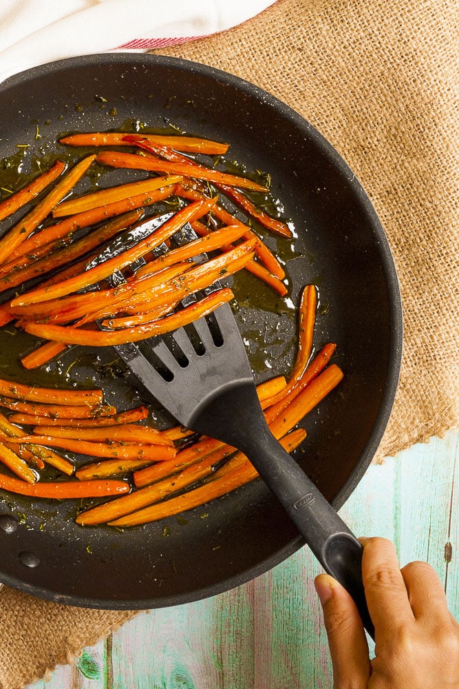 Black frying pan from above with thin carrot sticks in a herby oil. A hand with a slotted turner is taking some from the middle