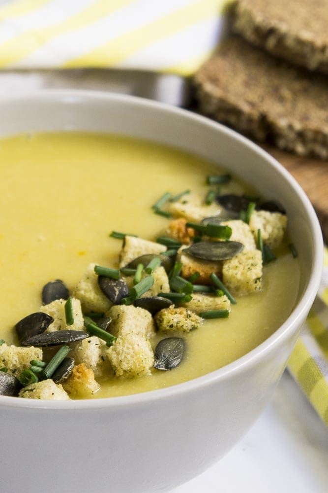 A bowl of yellow soup topped with croutons, pumpkin seeds and chives