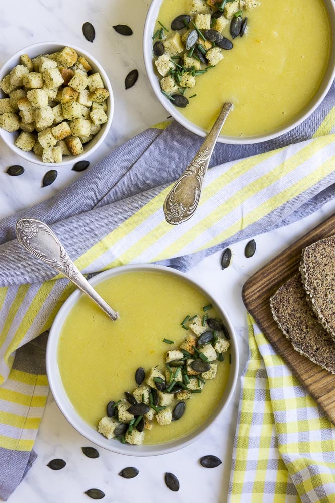 2 white bowls of yellow soup with spoons topped with croutons, pumpkin seeds and chives