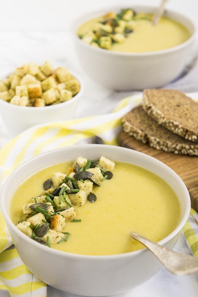 2 bowls of yellow soup with spoon topped with croutons, pumpkin seeds and chives