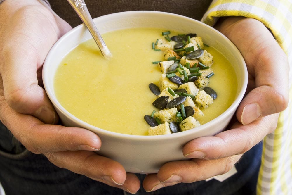 Two hands are holding a white bowl with yellow cream soup topped with croutons, pumpkin seeds and chopped chives. 