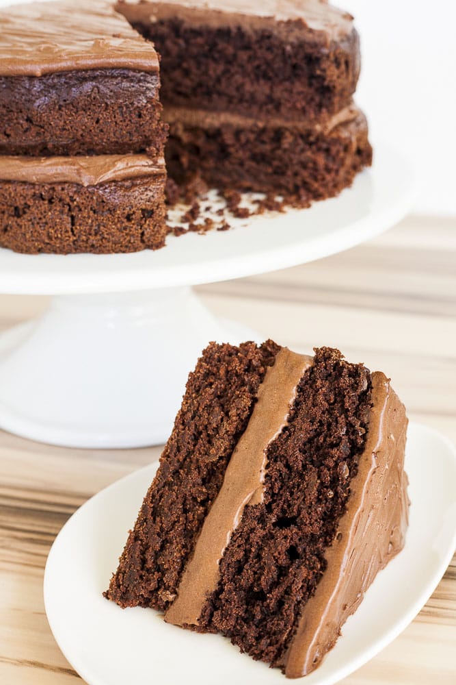 Vegan gluten-free cake at the back on a white cake stand. In front of it is a slice of cake on a white plate with a fork