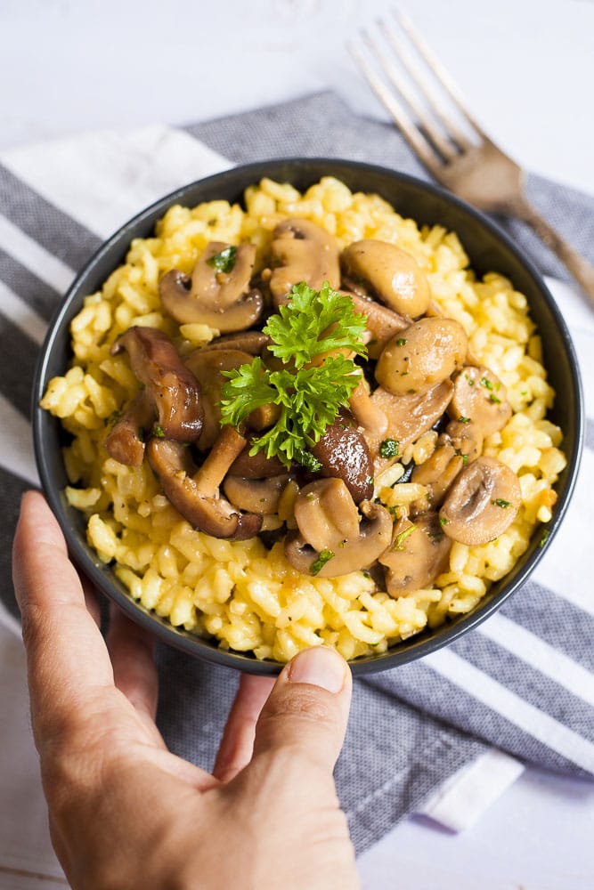 A hand is holding a black bowl with yellow rice topped with mushroom slices and chopped parsley