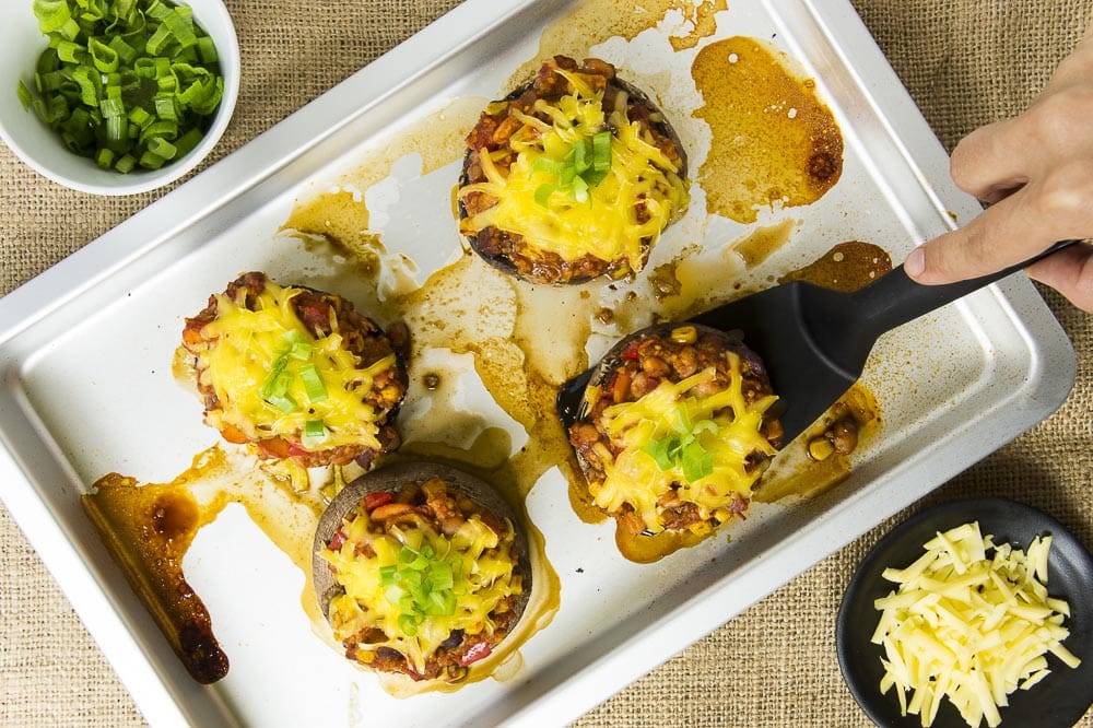 4 stuffed portobello mushrooms on the baking sheet with small bowls of grated cheese and chopped spring onion on the sides. 