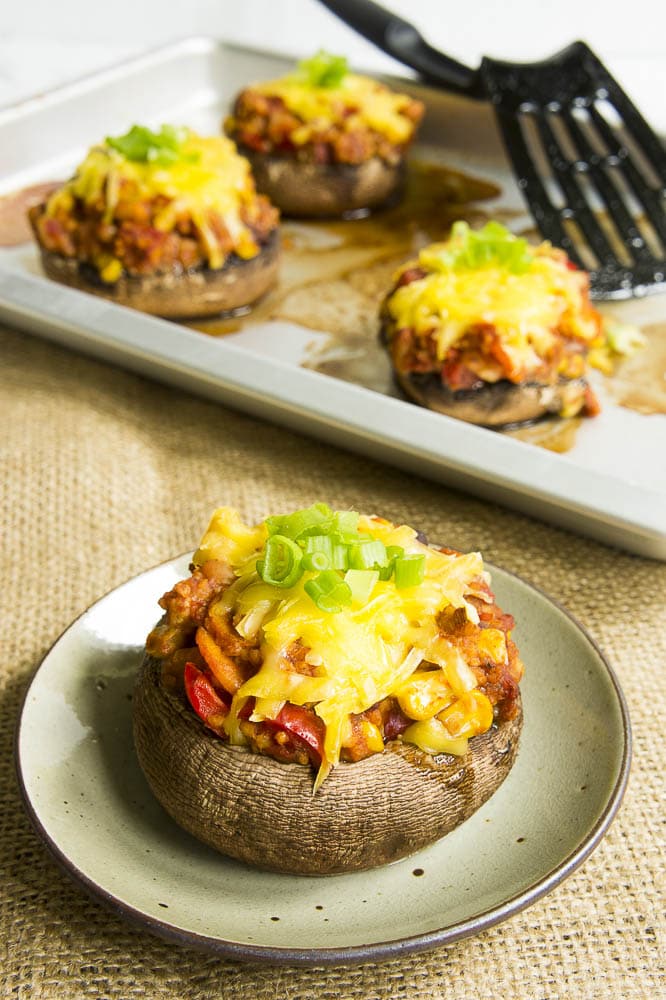 Small green plate with a large mushroom cap filled with chili topped with melted cheese and chopped spring onion. More stuffed mushrooms at the background on a silver baking tray