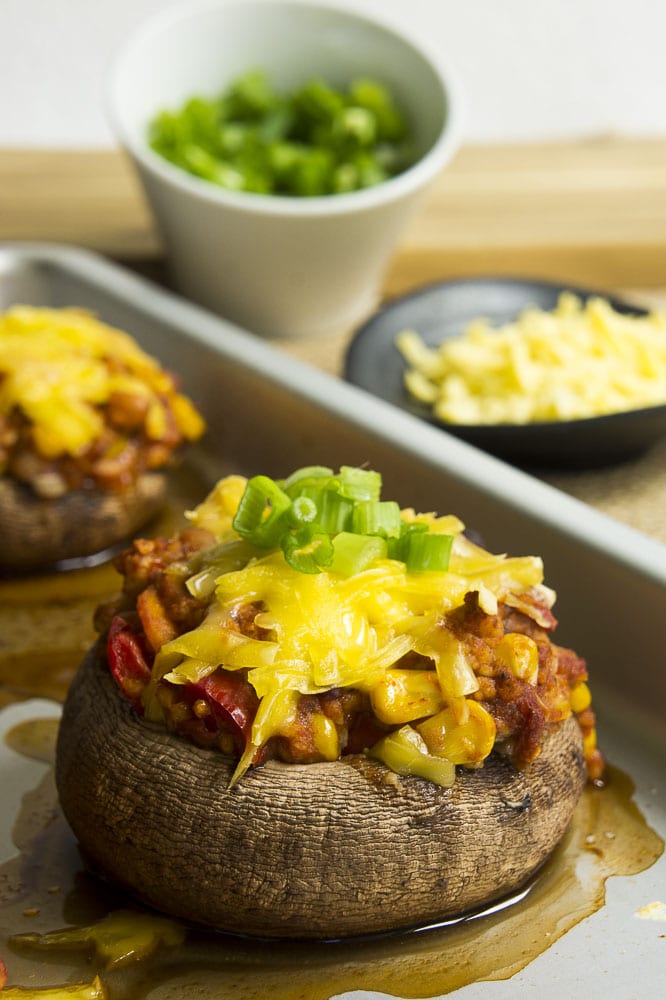 Small green plate with a large mushroom cap filled with chili topped with melted cheese and chopped spring onion. More stuffed mushrooms at the background on a silver baking tray