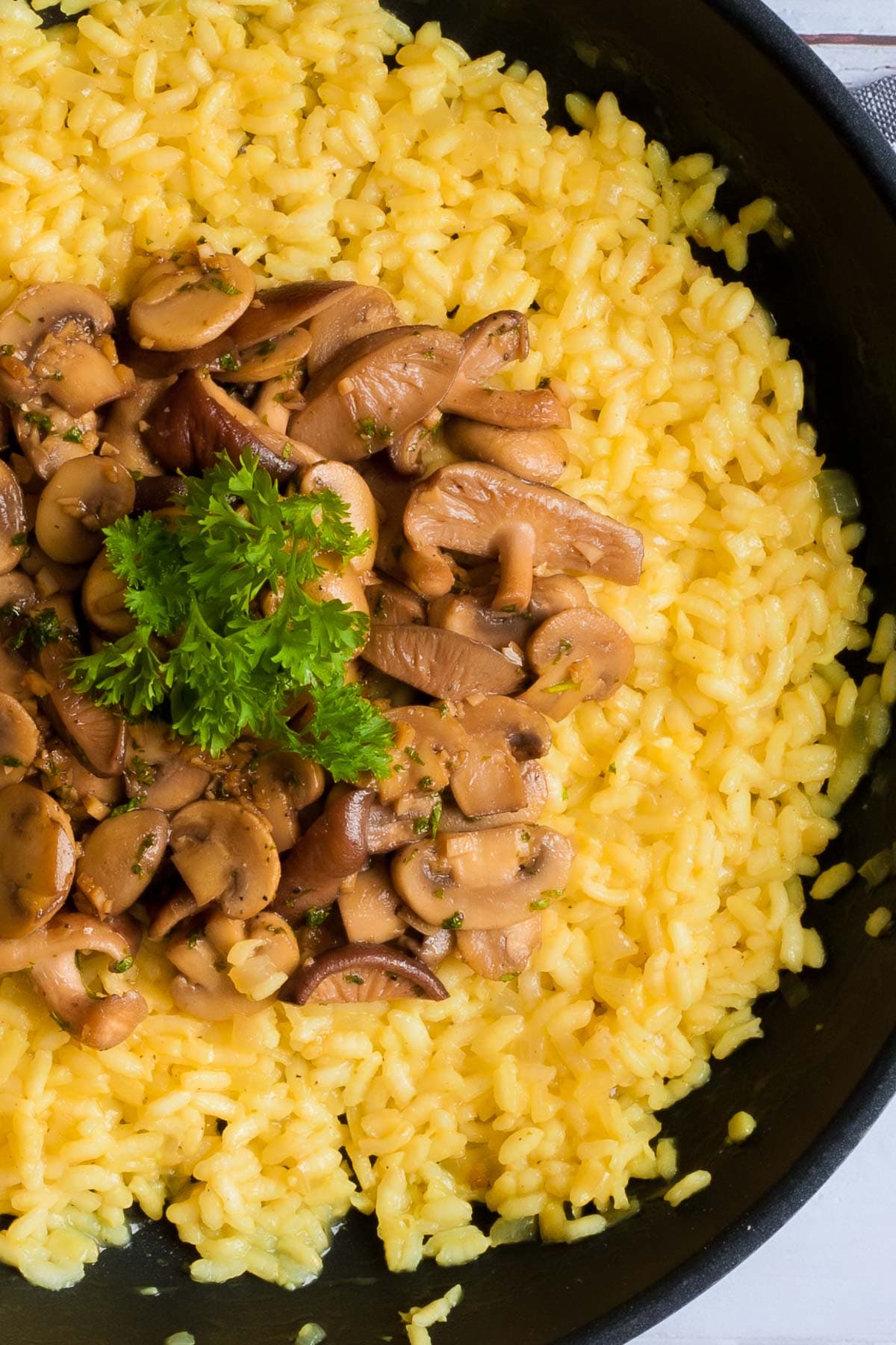 Large black frying pan with yellow creamy rice topped with brown mushroom slices and parsley leaves. 