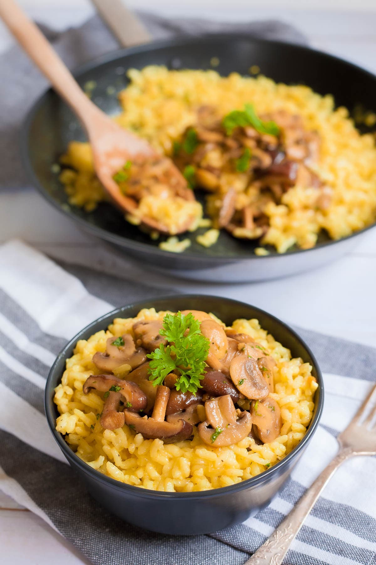 A black bowl of yellow creamy rice topped with brown mushroom slices and green fresh parsley. Leftover risotto is in a black frying pan at the back.
