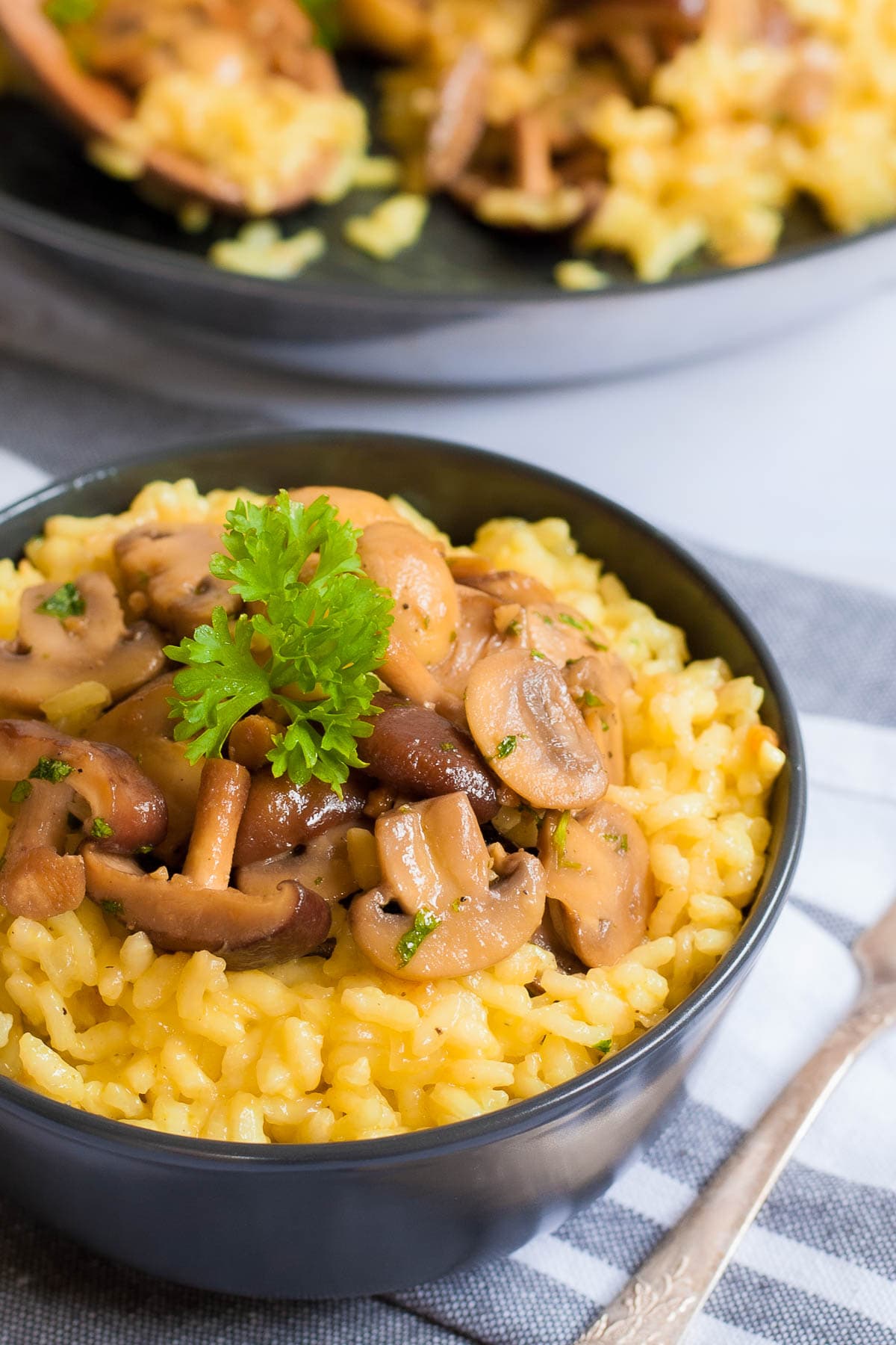 A black bowl of yellow creamy rice topped with brown mushroom slices and green fresh parsley. Leftover risotto is in a black frying pan at the back.