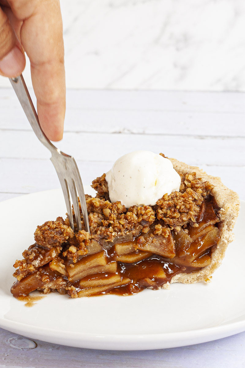 A slice of apple pie on a white plate with brown sauce, apple slices, crumbly topping and a scoop of white vanilla ice cream. There is a fork taking the first bite.