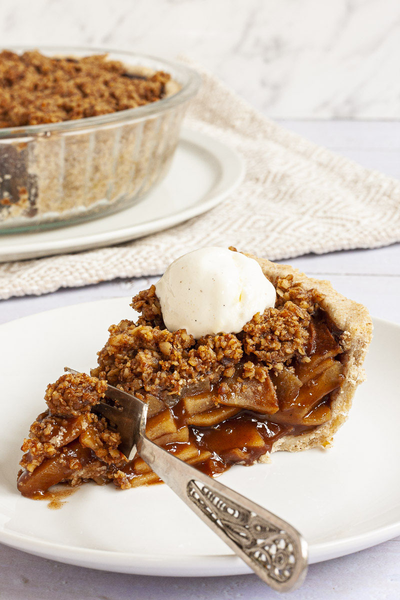 A slice of apple pie on a white plate with brown sauce, apple slices and crumbly topping. There is a fork taking the first bite.