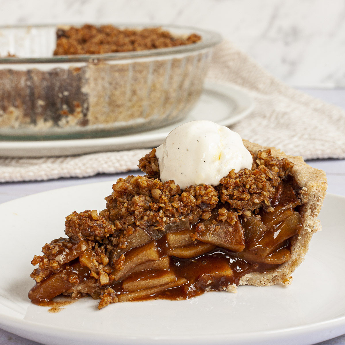 A slice of apple pie on a white plate with brown sauce, apple slices, crumbly topping and a scoop of white vanilla ice cream.