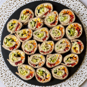 Lots of pinwheels with colorful veggies like orange carrots, red bell pepper, green cucumber, yellow corn in it placed on a black round plate with a white and gold border.