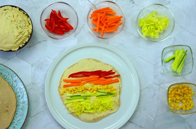 A white plate with a tortilla in the middle spread with a light brown sauce and colorful veggies like orange carrots, red bell pepper, green cucumber, yellow corn. The remaining ingredients are around it in glass bowls