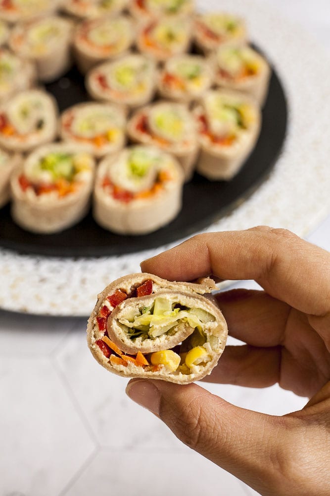 Lots of pinwheels with colorful veggies like orange carrots, red bell pepper, green cucumber, yellow corn in it placed on a black round plate with a white and gold border. A hand is holding one pinwheel