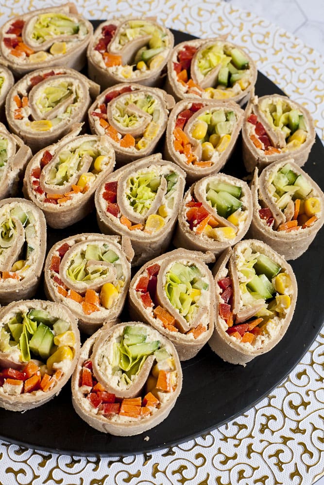 Lots of pinwheels with colorful veggies like orange carrots, red bell pepper, green cucumber, yellow corn in it placed on a black round plate with a white and gold border.