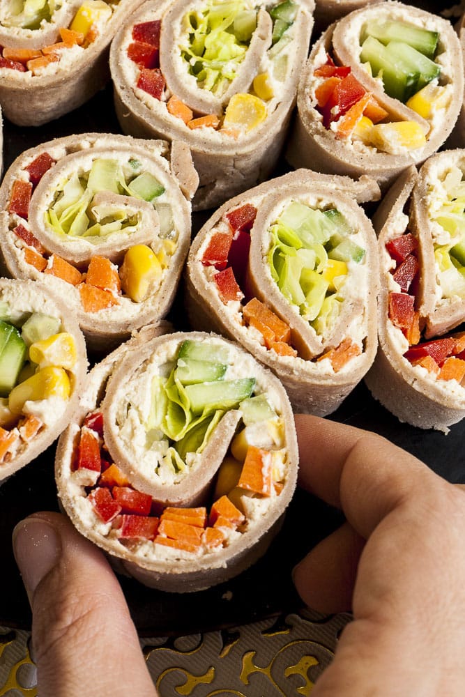 Lots of pinwheels with colorful veggies like orange carrots, red bell pepper, green cucumber, yellow corn in it placed on a black round plate with a white and gold border. A hand is about to grab one. 