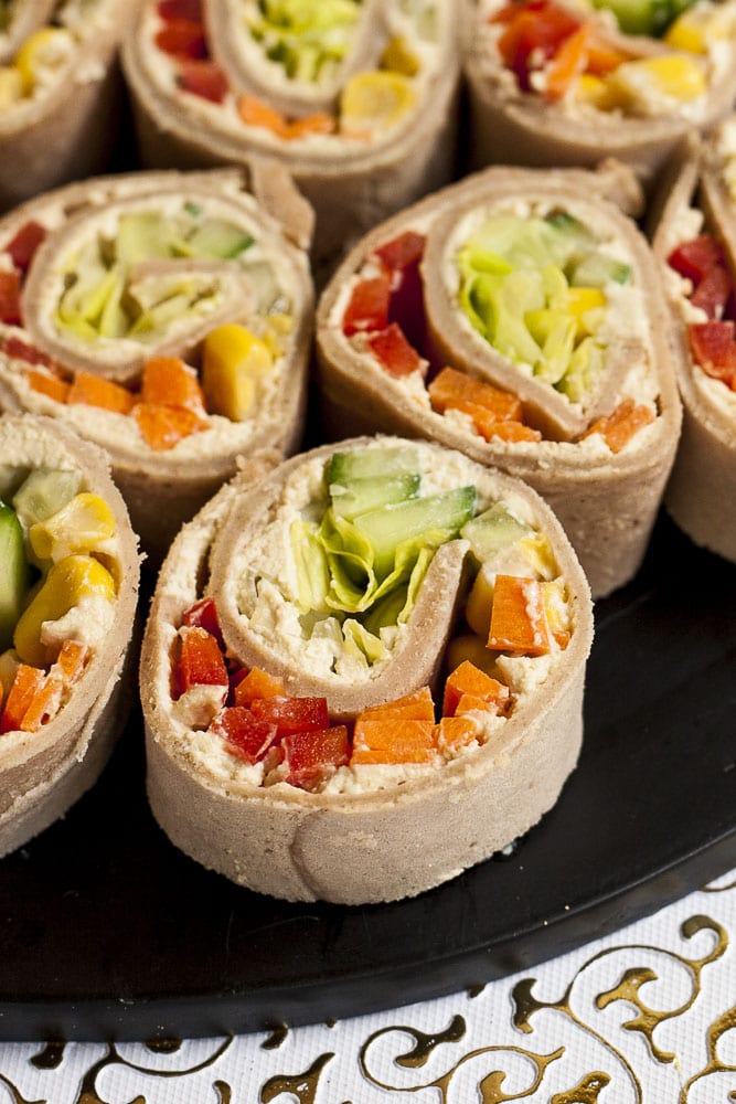 Lots of pinwheels with colorful veggies like orange carrots, red bell pepper, green cucumber, yellow corn in it placed on a black round plate with a white and gold border.