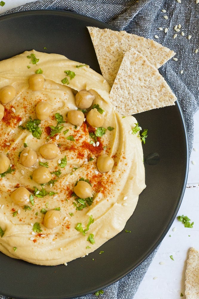 Black plate on grey tablecloth with oil-free hummus spread in the middle sprinkled with cooked chickpeas, fresh parsley, sweet paprika and sesame seeds. Flatbread triangle is just dipped into it.