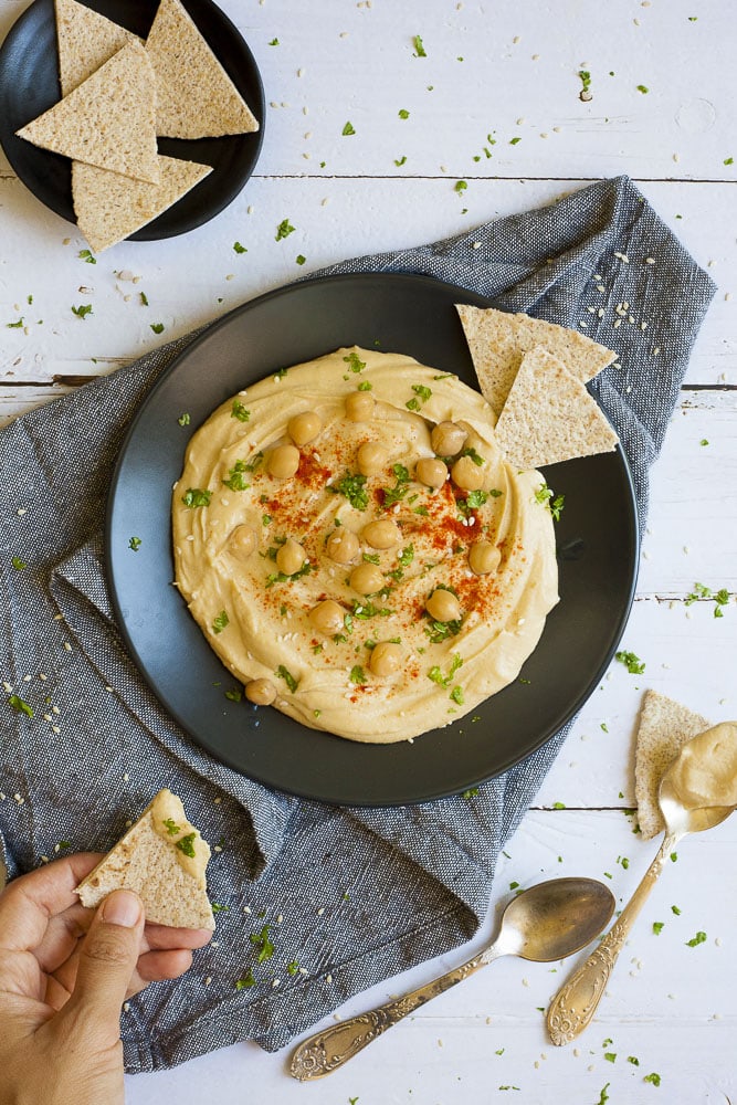 Black plate on grey tablecloth with oil-free hummus spread in the middle sprinkled with cooked chickpeas, fresh parsley, sweet paprika and sesame seeds. 