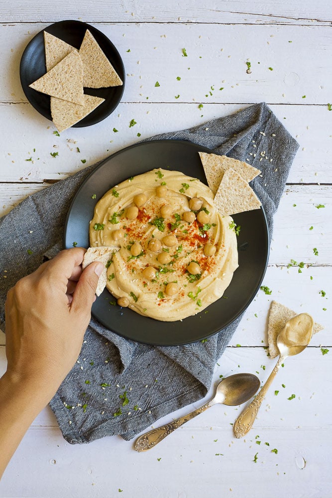 Black plate on grey tablecloth with oil-free hummus spread in the middle sprinkled with cooked chickpeas, fresh parsley, sweet paprika and sesame seeds. Flatbread triangle is just dipped into it.