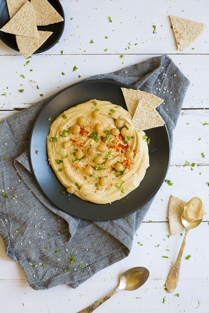 Black plate on grey tablecloth with oil-free hummus spread in the middle sprinkled with cooked chickpeas, fresh parsley, sweet paprika and sesame seeds. Flatbread triangles scattered around. 