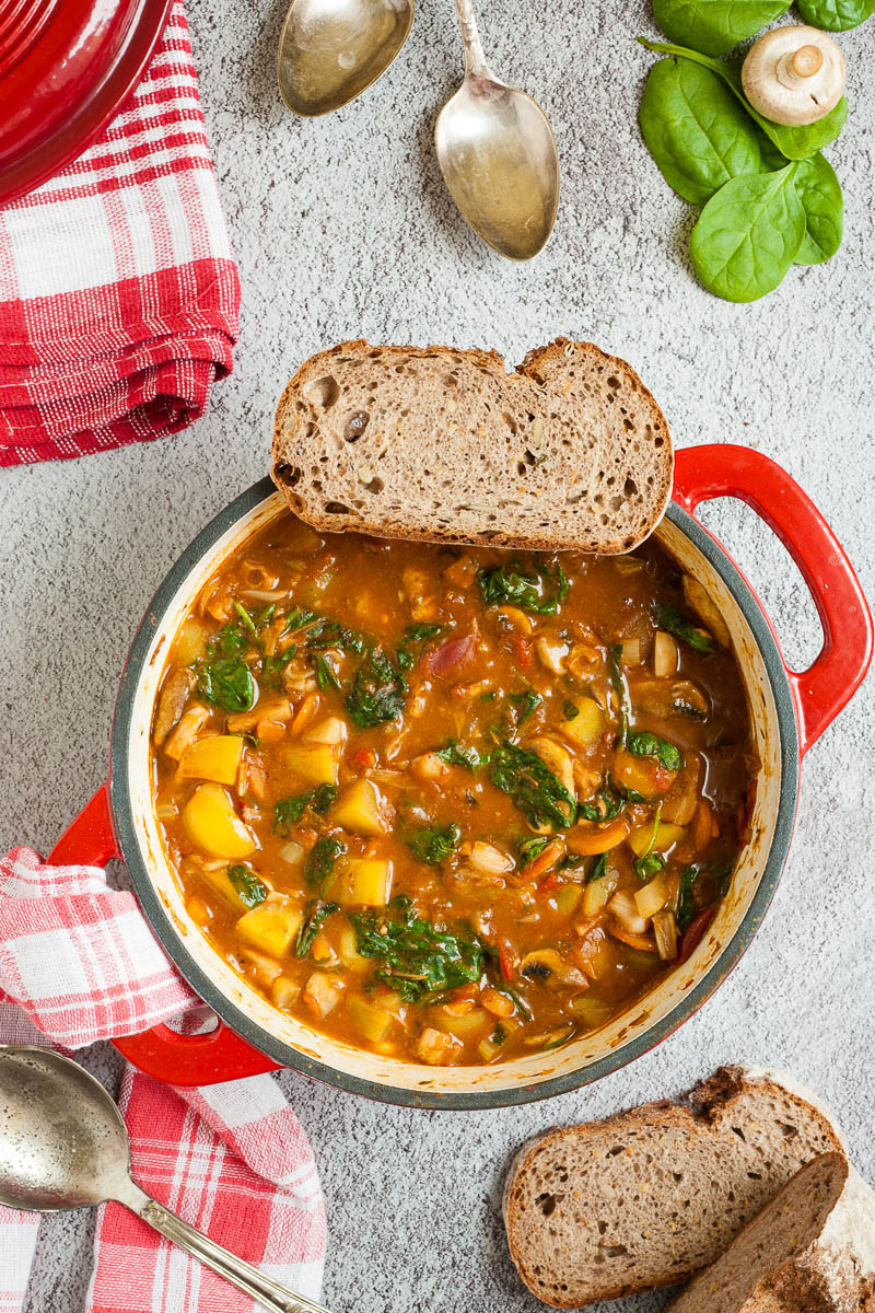 Red Dutch oven from above with a stew where you can see chopped potatoes, spinach leaves, carrot slices, tomatoes, and mushroom slices. A slice of bread is place on the rim. Red white table clothes are around with slices of bread, spinach leaves and spoons