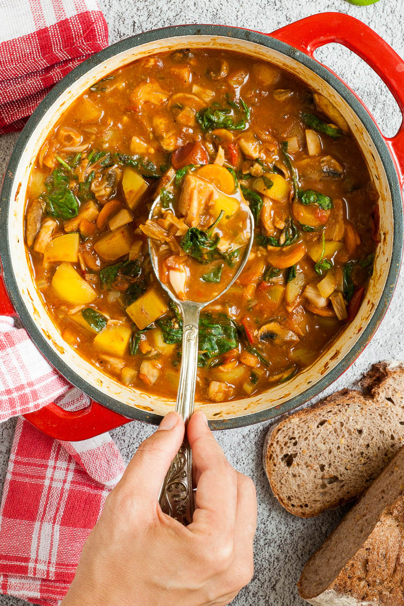 Red Dutch oven from above with a stew where you can see chopped potatoes, spinach leaves, carrot slices, tomatoes, and mushroom slices. Slices of bread is next to it. A hand is holding a spoon taking some from the middle