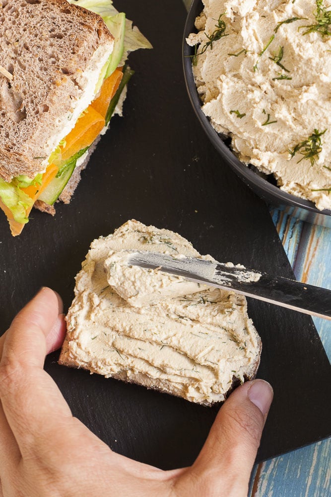 A slice of bread with tofu cream cheese. Next to it another finished sandwich and a bowl with the cream cheese