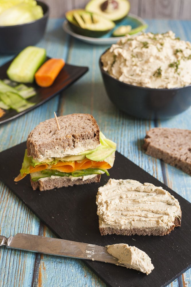 A slice of bread with tofu cream cheese. Next to it another finished sandwich. In the background a bowl with the cream cheese and other veggies to make a sandwich