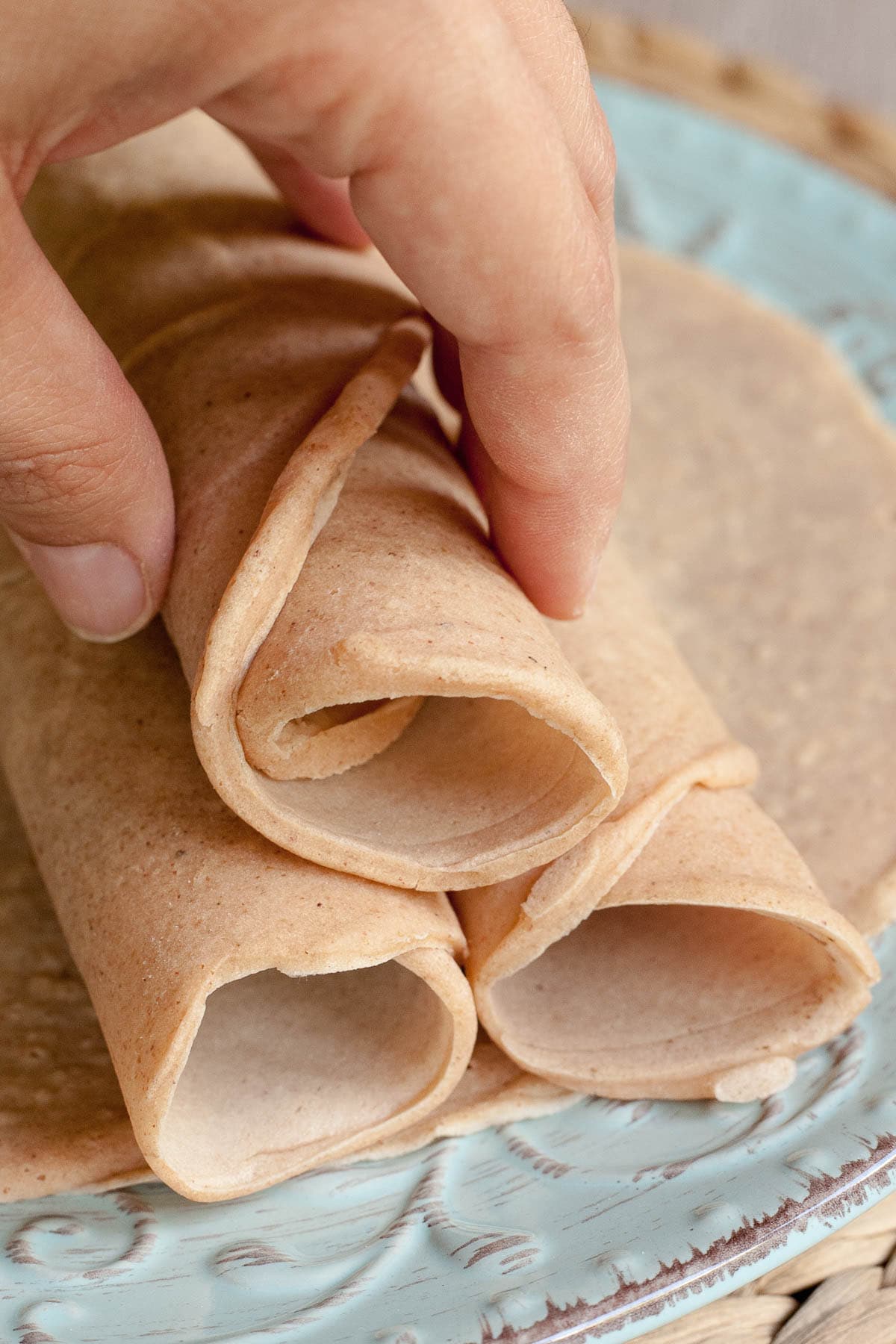 3 rolled tortillas on top of one flat placed on a light blue plate.A hand is holding the top one. 