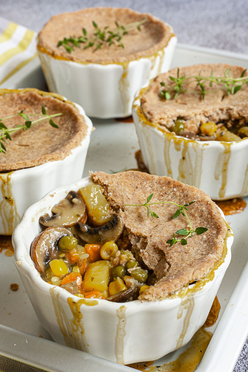 4 small white pots covered with brown pie crust and topped with green herbs. 2 of them has some crust missing and you can see the veggie stew within the yellow, green, orange, brown veggies of corn, green peas, mushrooms and carrots