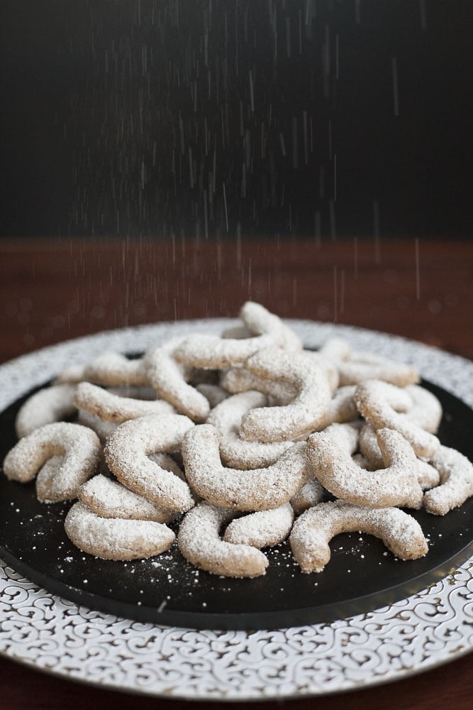 tack of walnut crescent cookies on a black plate and powdered sugar is falling from above