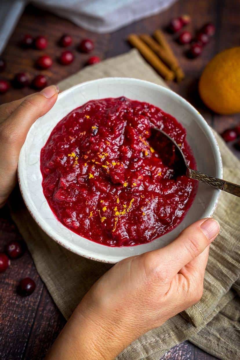 A hand is holding a small white bowl with a red-purple sauce sprinkled with orange peel. 