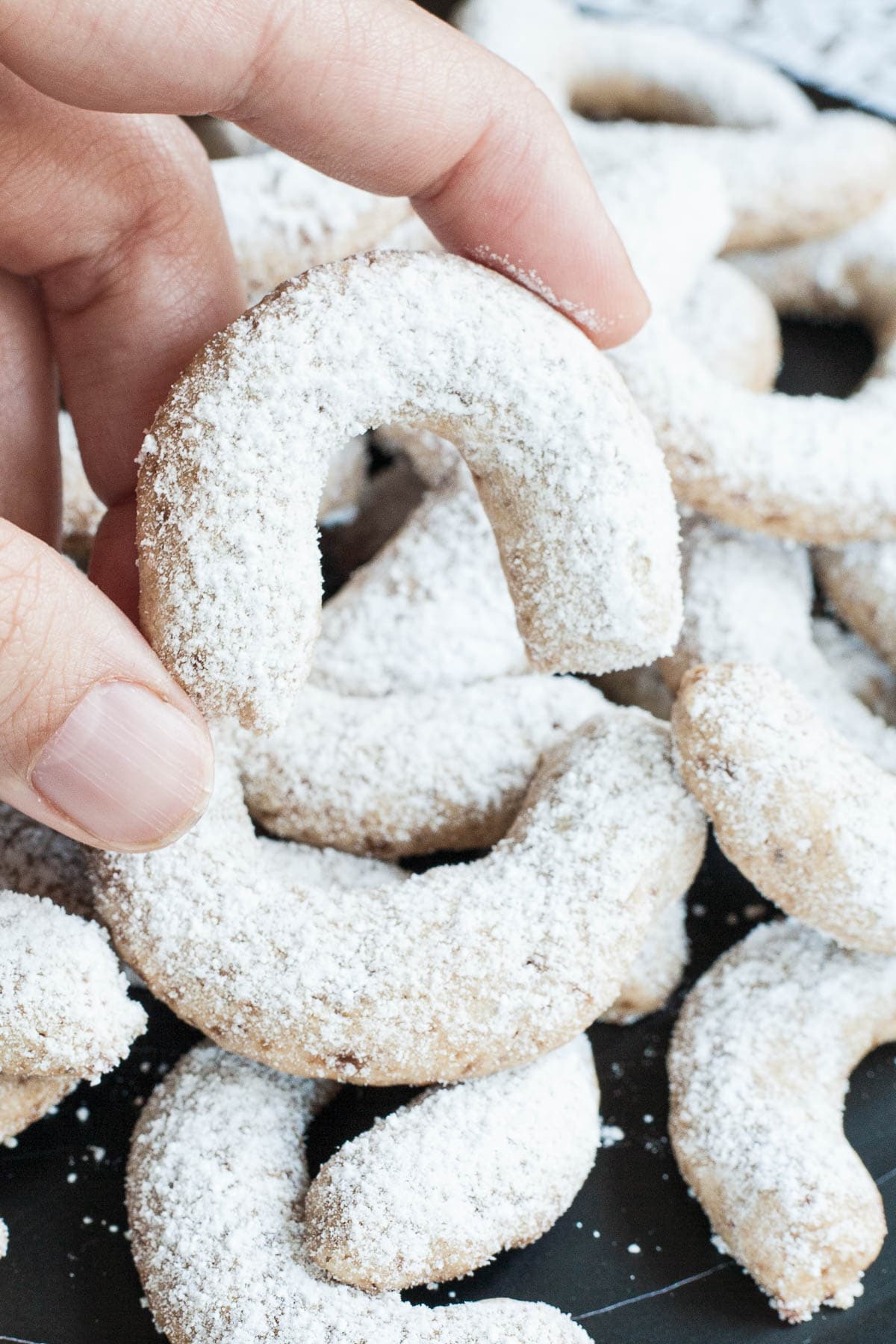 A large stack of crescent cookies on a black plate. Dusted with sugar. A hand is holding one cookie.