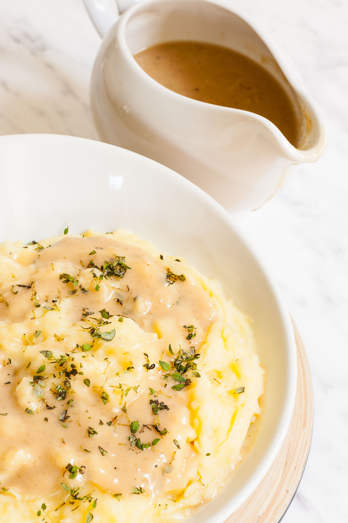 White bowl with mashed potatoes, with light brown gravy and chopped herbs. A gravy boat with the remaining gravy is next to it.