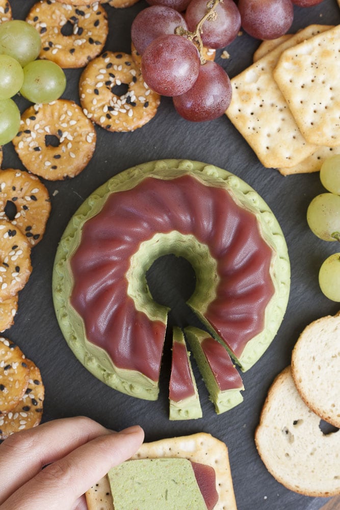 Small green bundt with purple cranberry jelly on top. It is served on a black plate with crackers and grapes. 