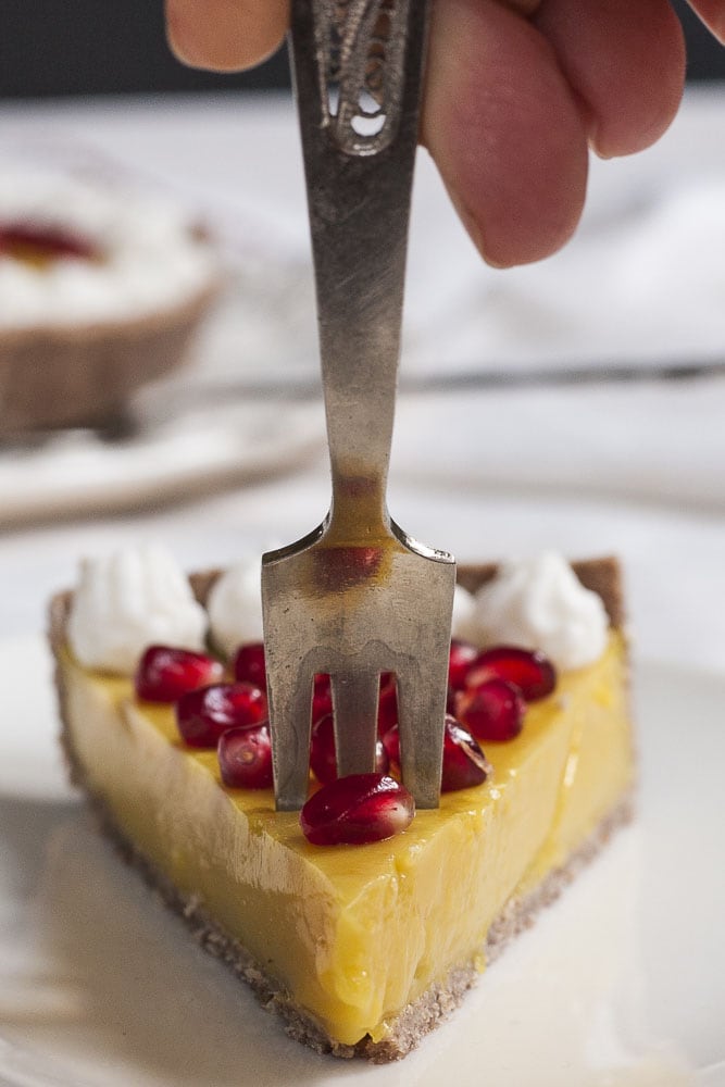 A slice of yellow custard tart is pierced with a fork from above. It is topped with white whipped cream and pomegranate seeds. 