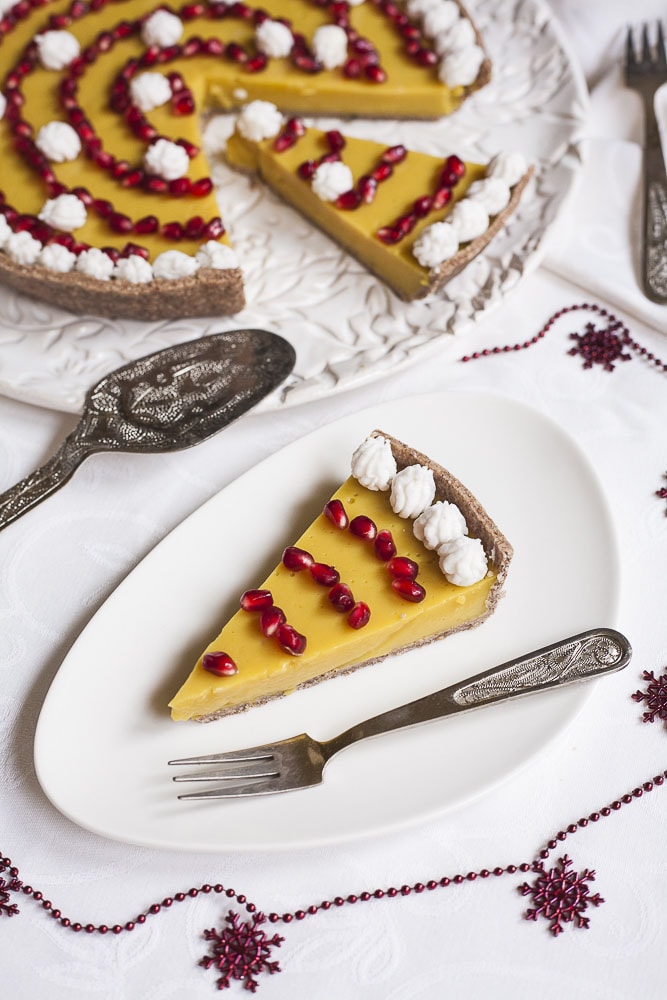 Slice of yellow custard tart on a white plate topped with white whipped cream and pomegranate seeds, the remaining tart is at the back on a white serving plate.
