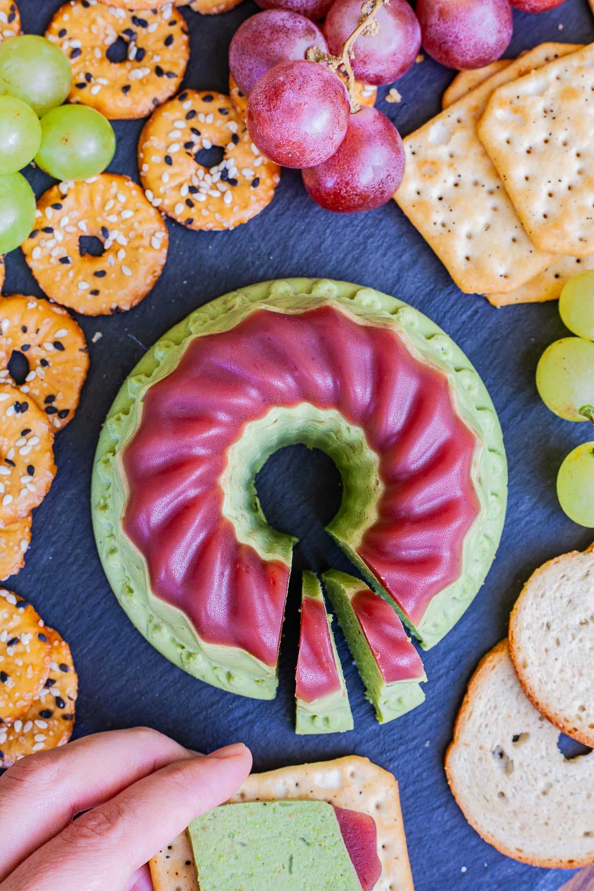 Green cheese shaped as a bundt topped with dark pink jelly. It is surrounded by grapes and different crackers. A hand is holding one cracker in the front with one slice of green cheese on it. 
